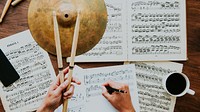 Hands holding drumsticks over sheet music, writing notes. Musical composition, sheet music, and drumsticks on a wooden table. Coffee cup nearby. Musician composing a song with drums.