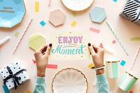 Hands holding a card with 'Enjoy every moment' surrounded by colorful party decorations, gifts, and plates. Celebration and joy with festive colors and patterns. Positive greeting card top view.