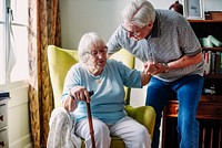 Elderly couple in a cozy room. An elderly man helps an elderly woman with a cane. The elderly couple shares a moment in a warm, inviting space. Senior man is helping woman up from chair.