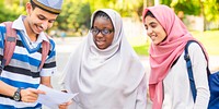 Three young adults in casual attire, smiling and looking at a paper. They wear backpacks and headscarves, enjoying a sunny day outdoors together. Happy international students outside of school.