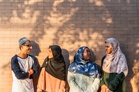 Group of four muslim teenagers standing against a brick wall, wearing hijabs and casual clothing, enjoying a sunny day. Smiling, chatting, and relaxed atmosphere. Muslim teenage students hanging out.