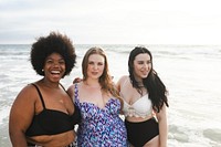 Three women in swimsuits enjoying the beach. They are smiling and standing by ocean. Women representing different ethnicities and body types. Diverse women at beach, body positivity and inclusivity.