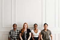 Group of four people sitting against a white wall, smiling. Diverse group includes men and women, showcasing diversity and unity in a casual setting. Happy diverse people sitting in a row.