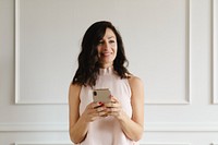 Smiling woman holding a smartphone, standing against a white wall. Casual attire, relaxed pose, smartphone in hand, modern technology, cheerful expression. Content creator on social media.