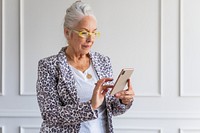 Elderly woman with white hair and glasses using a smartphone. She wears a leopard print jacket. The woman is focused on her phone, standing indoors. Senior businesswoman working on phone.