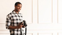 Man holding a camera, wearing a plaid shirt, smiling. Photography enthusiast, casual style, indoor setting. Capturing moments, enjoying photography. African American man in minimal modern room.