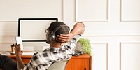 Man relaxing with headphones, smartphone in hand, sitting at desk. Casual setting with computer. Relaxing and using smartphone in office. African American man relaxing, using smartphone by desk.