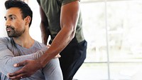 A person assists another in a stretching exercise. Focus on arm movement and physical support. Indoor setting with natural light. Fitness and wellness theme. Workout exercise concept photography.