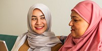 Two women in hijabs smiling, one in gray, one in pink. Women are sitting together, sharing a moment. Hijabs, smiles, and togetherness are key themes. Happy warm muslim family photo.