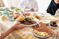 A diverse group shares a meal with lamb chops, bread, and dates. Hands pass dishes around a table filled with Middle Eastern cuisine, fostering community and togetherness. Muslim family dinner.