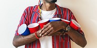 Man holding red and blue cups, wearing a red striped shirt. Red and blue cups, casual style. Red and blue cups, party vibe. Red and blue cups, fun atmosphere. Drunk African American man at a party.