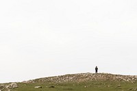 A solitary figure stands on a rocky hilltop under a vast, cloudy sky. The lone person on the hill top gazes at the expansive sky, embracing solitude and nature. Man hiking on mountain background.