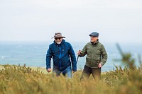 Two older men hiking outdoors, wearing jackets and hats. They enjoy nature, walking through fields. Hiking and nature are key themes in this outdoor scene. Happy seniors hiking in British nature.