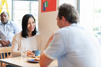 Two people at a cafe table, smiling and talking. Woman with long black hair, man with short hair. Casual setting, coffee cups on the table, relaxed atmosphere. Woman and man chatting in cafe.