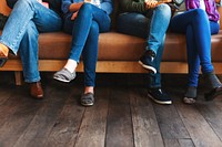 Four people sitting on a couch, legs crossed, wearing blue jeans and casual shoes. Wooden floor, casual setting, diverse footwear styles. People sitting in a row on a sofa wearing jeans and denim.