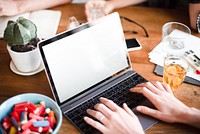 Hands typing on a laptop at a wooden table with a cactus, a bowl of capsules, and a glass of juice. Laptop, typing, and workspace elements visible. Blank laptop screen on messy desk.