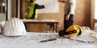 Construction site with blueprints, hard hat, and safety gear. Focus on planning and safety. Workers in background, blurred, discussing project details. House renovate and construction concept.