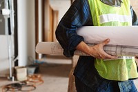 Person in construction gear holding blueprints. Construction worker with plans. Safety vest, blueprints, construction site. Focus on construction work. House renovate and construction concept.