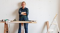 A woman stands confidently by a workbench with tools. She wears a plaid shirt and jeans. A ladder is nearby, suggesting a DIY or construction project. Woodworking and construction work concept.