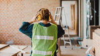 Female construction worker wearing safety gear, including a vest and headphones, in a building site. Worker focuses on safety, surrounded by construction tools and materials. Construction in a home.