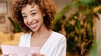 Smiling woman holding a paper, surrounded by plants. Curly hair, casual white shirt. Bright smile, cheerful expression. Indoor setting, relaxed atmosphere. Woman in cozy room portrait.