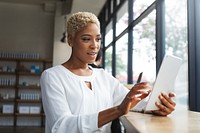 Woman using tablet in bright cafe. African American woman focused on tablet. Casual setting, woman in white shirt, using technology, enjoying her time. Casual business woman in cafe portrait.