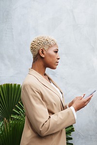 Profile of a woman in a beige blazer holding a phone, standing by green plants against a gray wall. Short curly hair, focused expression, modern style. Businesswoman in business formal attire.