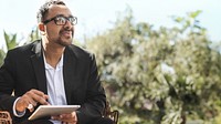 Man in a suit with glasses holding a tablet outdoors. Businessman with tablet, wearing glasses, sitting outside. Professional male with tablet in nature. A man using a tablet outdoor.