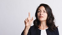 Woman gesturing with finger, focused expression. Brunette woman, pointing, wearing a dark blazer. Confident woman, making a point, isolated background. Woman gesturing against white wall.