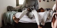 Person in cozy sweater and sweatpants sits on bed with tablet. Casual, relaxed setting with clothes and teddy bear. Comfortable, informal atmosphere. Teenager in a bedroom.