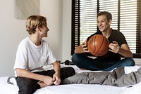 Two young males sitting on a bed, one holding a basketball. Casual setting with natural light. Relaxed atmosphere, friendly interaction, basketball focus. Caucasian brothers photo.