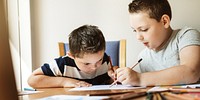 Two young boys drawing together at a table. One boy helps the other with a pencil. Creative activity, teamwork, and learning in a cozy setting. Children playing and writing in modern room.