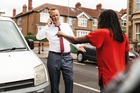 Man and woman in a street dispute. One in a suit, talking on the phone, calling car insurance, the other in a red shirt gesturing to a car in a neighborhood setting. Car accident concept.