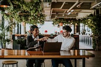 Two people in a modern office, engaged in a friendly fist bump. They sit at a wooden table with a laptop, surrounded by greenery and stylish decor. Diverse colleagues working together.