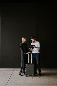 Two people with luggage, one holding a tablet. Business attire, discussing travel plans. Indoor setting, dark background, professional atmosphere. Professional traveling for business.