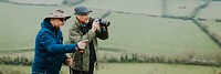 Two older men outdoors, one with a camera, enjoying nature. Both wear hats and jackets, standing in a green field, capturing the scenic landscape. Two elderly people hiking on hill, outdoor scene.