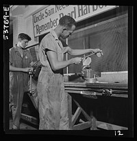 NYA (National Youth Administration) work center, Brooklyn, New York. Two bench workers, one white and one a , who are receiving training in machine shop practice, removing the burrs from a side piece for a metal pulley block. Sourced from the Library of Congress.