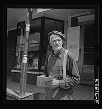 West Danville, Vermont. Frank Goss, seventy-one year old farmer, in front of Gilbert S. Hastings General Store and post office reading his mail which includes a card saying that his last year's hired man "wont be aroun for hayin' this year on account of he's in Californi' in the Navy". Sourced from the Library of Congress.