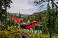 A girl with blonde hair sits in a red and gray hammock overlooking mountains, trees, and water.. Original public domain image from Wikimedia Commons