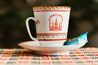 A ceramic cup with a castle on it sitting on a saucer with a biscuit in a wrapper. Original public domain image from <a href="https://commons.wikimedia.org/wiki/File:Kremlin_cup_of_tea_with_candy._(Unsplash).jpg" target="_blank" rel="noopener noreferrer nofollow">Wikimedia Commons</a>