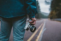 A man in denim clothing stands on an empty paved road holding a camera. Original public domain image from Wikimedia Commons