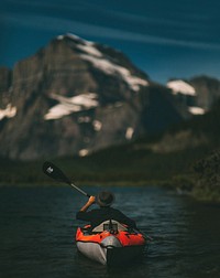 A man wearing a hat paddling in a blow up orange canoe in Swiftcurrent Lake. Original public domain image from Wikimedia Commons