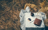 A pine cone, journal, leather bag, and reed diffuser on a lace tablecloth in a wheatfield in Thailand. Original public domain image from <a href="https://commons.wikimedia.org/wiki/File:Artsy_Tablescape_(Unsplash).jpg" target="_blank" rel="noopener noreferrer nofollow">Wikimedia Commons</a>