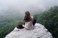 A lone woman sitting on a rocky ledge overlooking a forest with a red hat on her side. Original public domain image from Wikimedia Commons