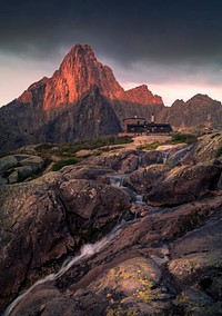 Modern house at the top of a rocky hill with mountains and a creek in the background and foreground during sunset. Original public domain image from Wikimedia Commons