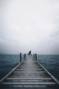 A person jumping in the air on the edge of a dock by a lake in Punta Saint Vigilio on a cloudy day. Original public domain image from Wikimedia Commons