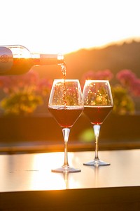 A person pouring red wine into one of two glasses with flowers and the setting sun in the background. Original public domain image from <a href="https://commons.wikimedia.org/wiki/File:Sunset_and_wine_(Unsplash).jpg" target="_blank" rel="noopener noreferrer nofollow">Wikimedia Commons</a>
