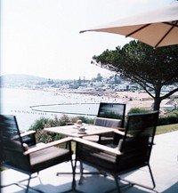 A patio with three chairs at a square table with a view on a crowded beach and a coastal town. Original public domain image from <a href="https://commons.wikimedia.org/wiki/File:Au_calme_(Unsplash).jpg" target="_blank" rel="noopener noreferrer nofollow">Wikimedia Commons</a>