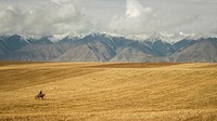 A person on horseback riding through a golden field at the foot of a snow-capped mountain range. Original public domain image from <a href="https://commons.wikimedia.org/wiki/File:Camping_on_the_shores_of_lake_Issyk_Kul_(Unsplash).jpg" target="_blank" rel="noopener noreferrer nofollow">Wikimedia Commons</a>