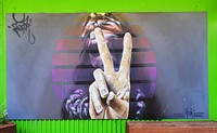 Another Wongi "Freak" Wilson Mural. Wongi’s work, the largest of the three, depicts a monotone female figure, with her hand thrust forward, two fingers raised in a peace sign, the hand bursting to life in colour and sharp detail. A segmented disc of translucent yellow, orange, pink and purple separates the figure and the hand, framing the image and popping against the dark grey background. Wongi noted that he had chosen the image to play with a “beachy vibe” in honour of the location, but without the usual cliché, instead of seagulls or surfers, his character has a summery, music festival feel.The image is another example of Wongi’s ever impressive photo realistic technique, highlighting his aerosol mastery, an expertise that was made apparent to the crowds that stopped and watched the Christchurch legend in action, gaining insight into the sketching and refining process with which he builds form and brings his images to life. Original public domain image from Flickr